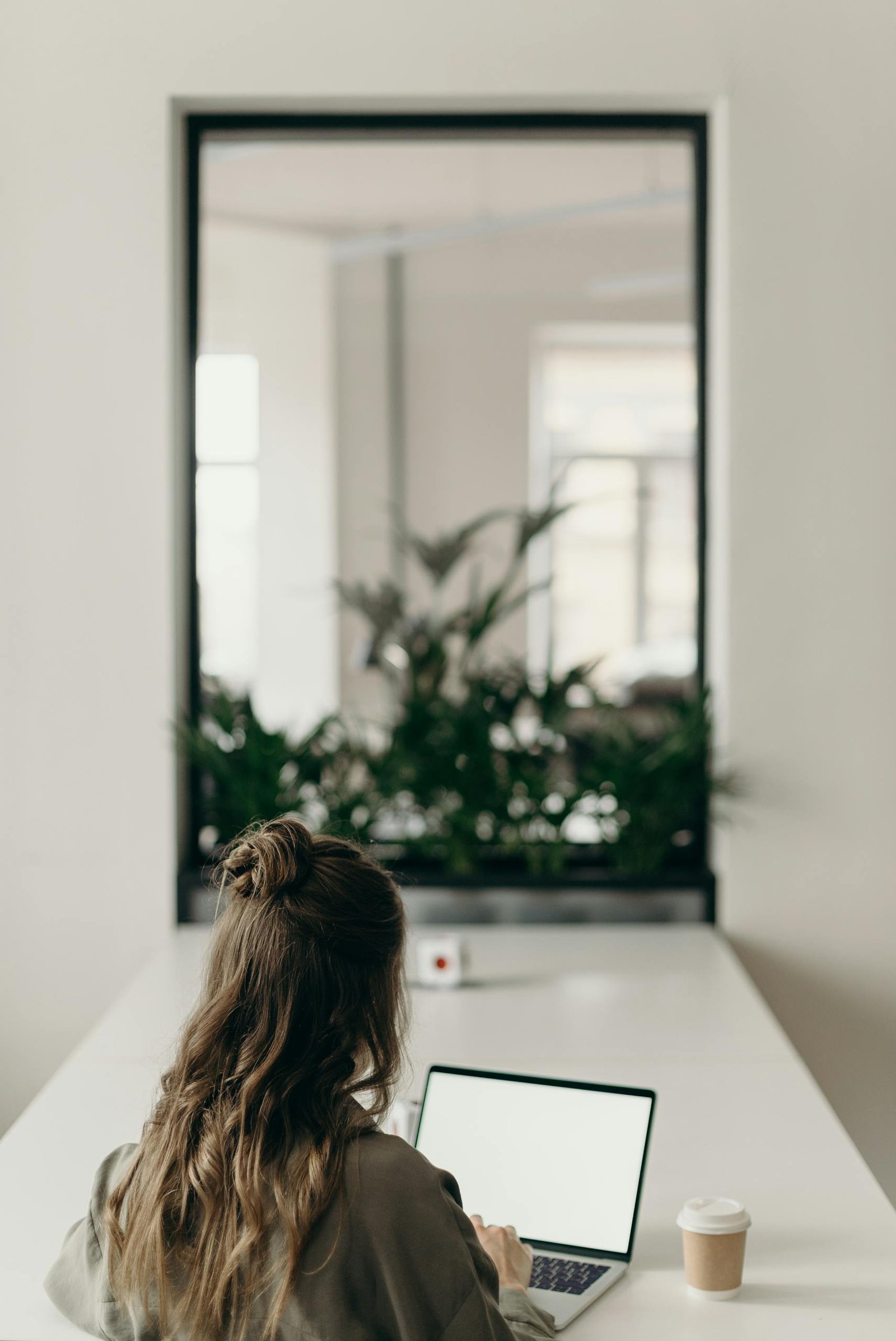 Deine ultimative Reiseplanung: Tipps, Tricks & ein bisschen Chaos A woman with curly hair works on a laptop, facing a window with plants, symbolizing remote work.